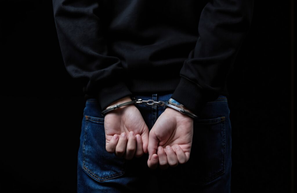 Teenage boy in black hoodie and jeans is arrested with hands cuffed behind his back. Dark studio background emphasizes his confinement. Represents crime, justice and consequence for youth.